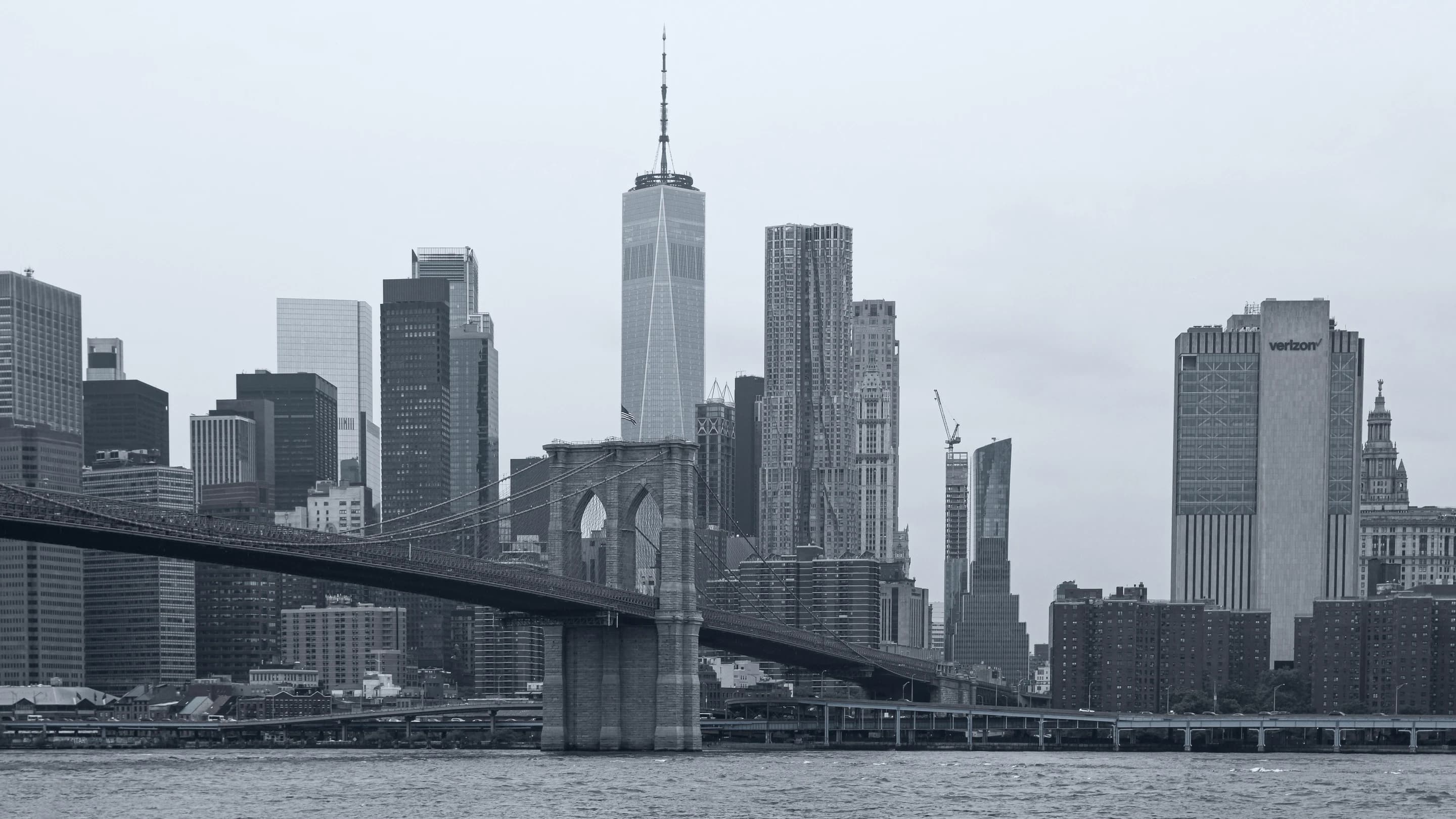 Black-and-white view of the Brooklyn Bridge and Lower Manhattan skyline, with One World Trade Center rising above the waterfront.