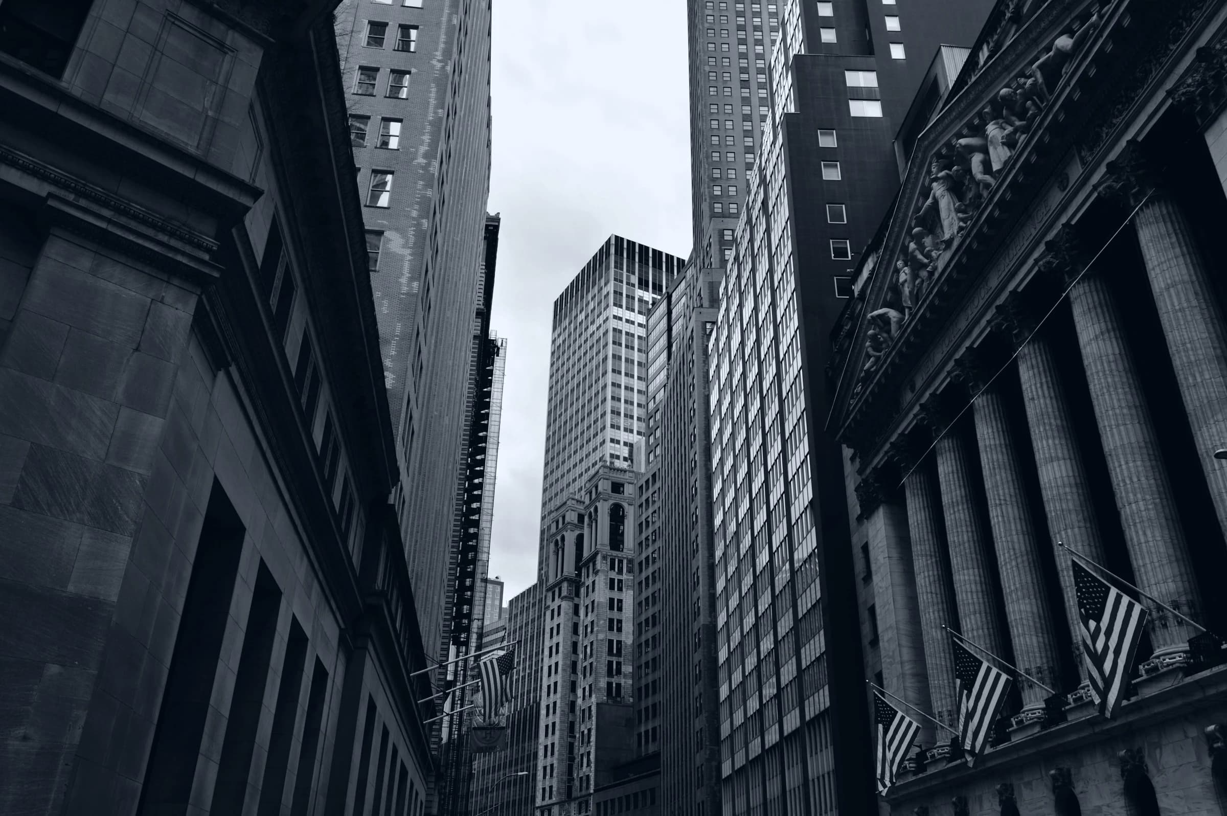 Black-and-white view looking up between Wall Street buildings, with the New York Stock Exchange facade and American flags.