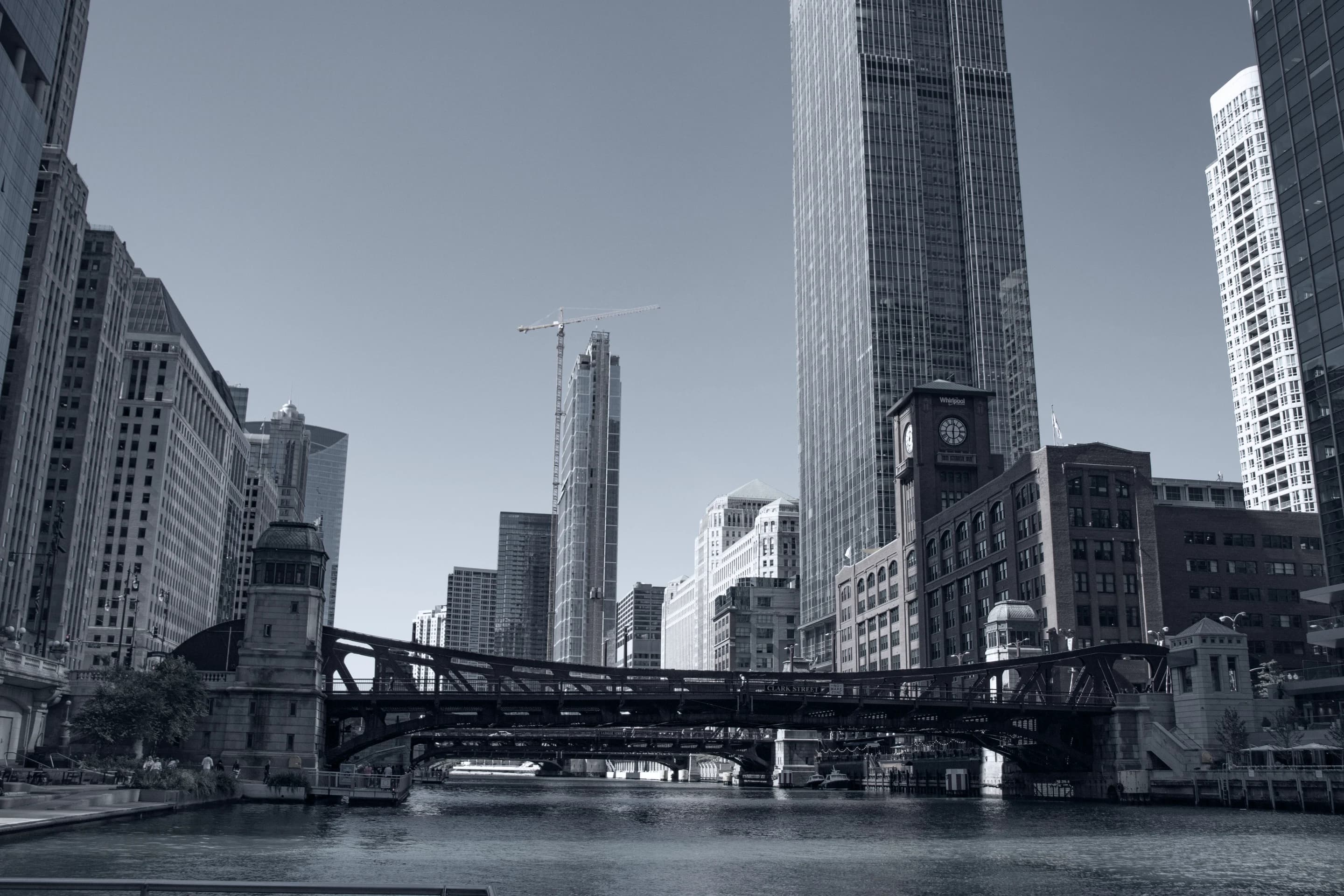 Black-and-white view along the Chicago River, framed by high-rise buildings and steel bridges beneath a clear sky.