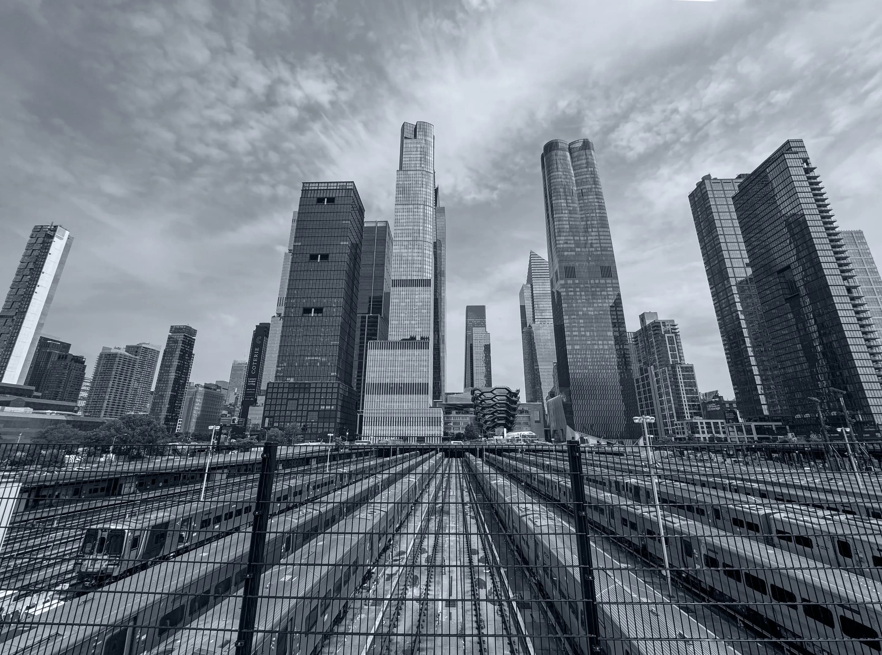 Black-and-white view over rail tracks towards the towers of Hudson Yards beneath a broad clouded sky.